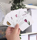 A hand holding a series of folded cards with poems written on them, each card has a different floral illustration at the bottom.