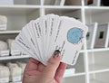 Hand holding a deck of cards with motivational phrases and a blue circle design, with shelves in the background.
